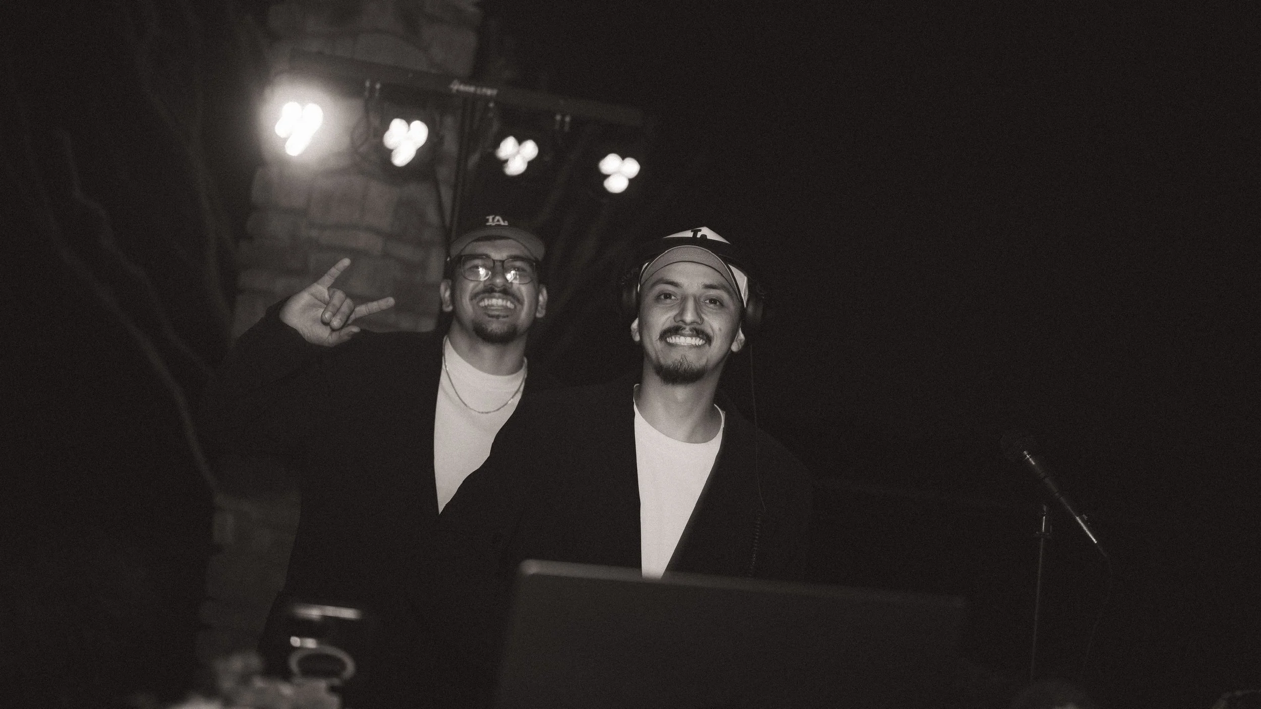 Two smiling men with glasses and hats, one making a rock sign with his hand, in a dark room with stage lights overhead.