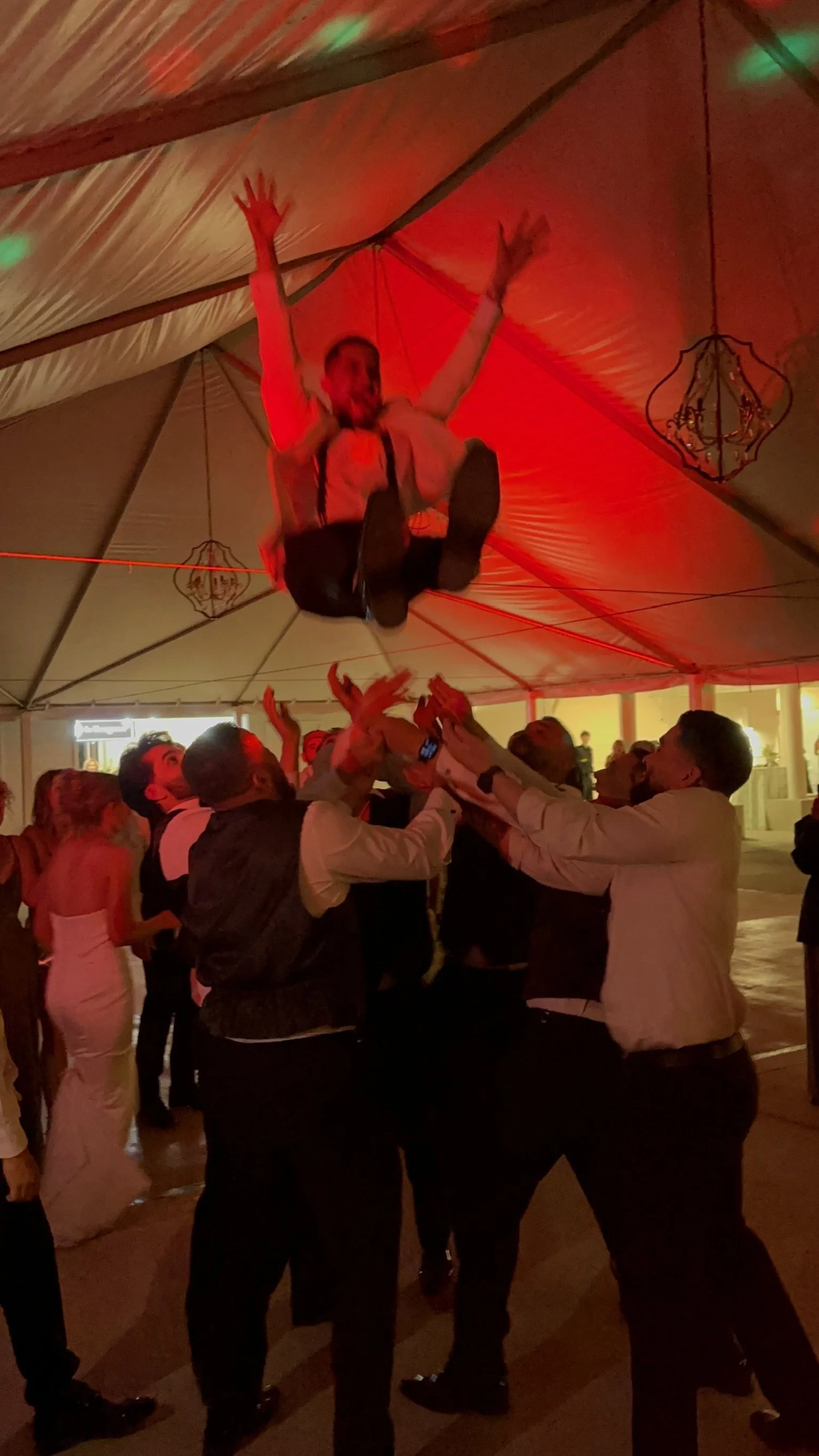 People celebrating at a wedding reception with a man being lifted into the air by friends under a decorated tent with hanging lanterns.