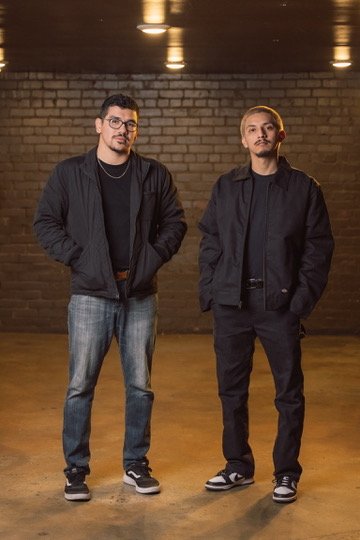 Two young men standing in a dimly lit indoor space with a brick wall background, dressed in dark casual clothing and sneakers.