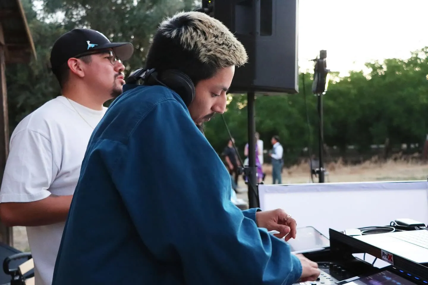 A DJ with headphones around his neck is operating a mixing console outdoors, with a man standing behind him and other people in the background.