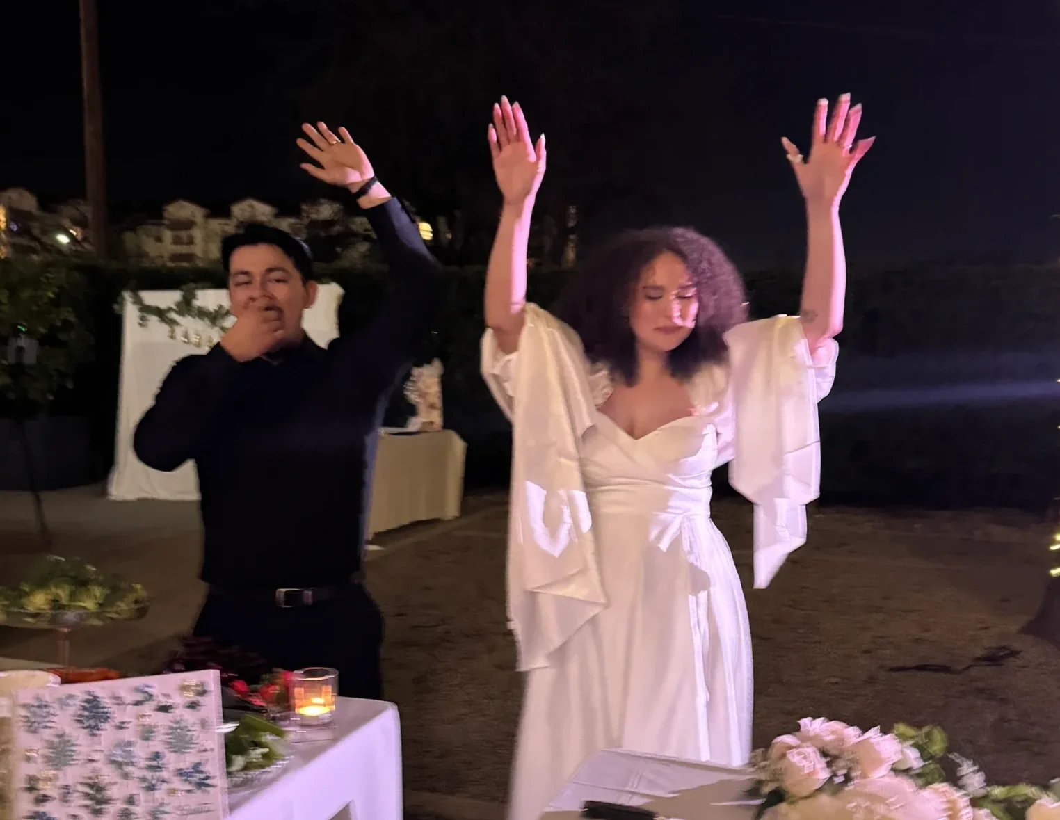 A man and a woman at a nighttime celebration, possibly a wedding. The man is wearing a black outfit and the woman is in a white dress with her arms raised. They are outdoors with tables, flowers, and candles.