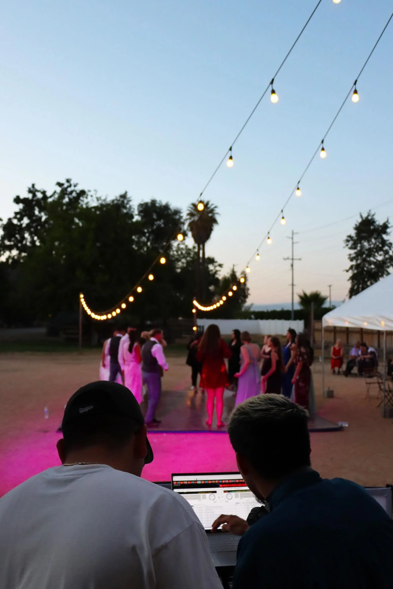 People gathering outdoors at dusk with string lights overhead, a tent, and trees in the background. Two individuals are working on a laptop in the foreground, possibly DJing or managing sound for the event.