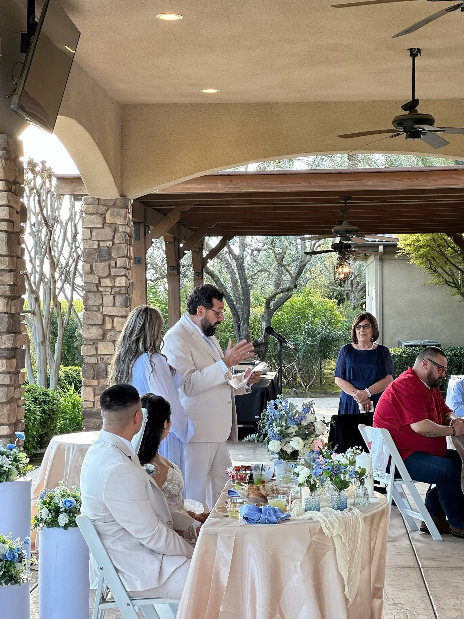 A wedding reception outdoor under a patio with guests seated at decorated tables and a woman giving a speech.
