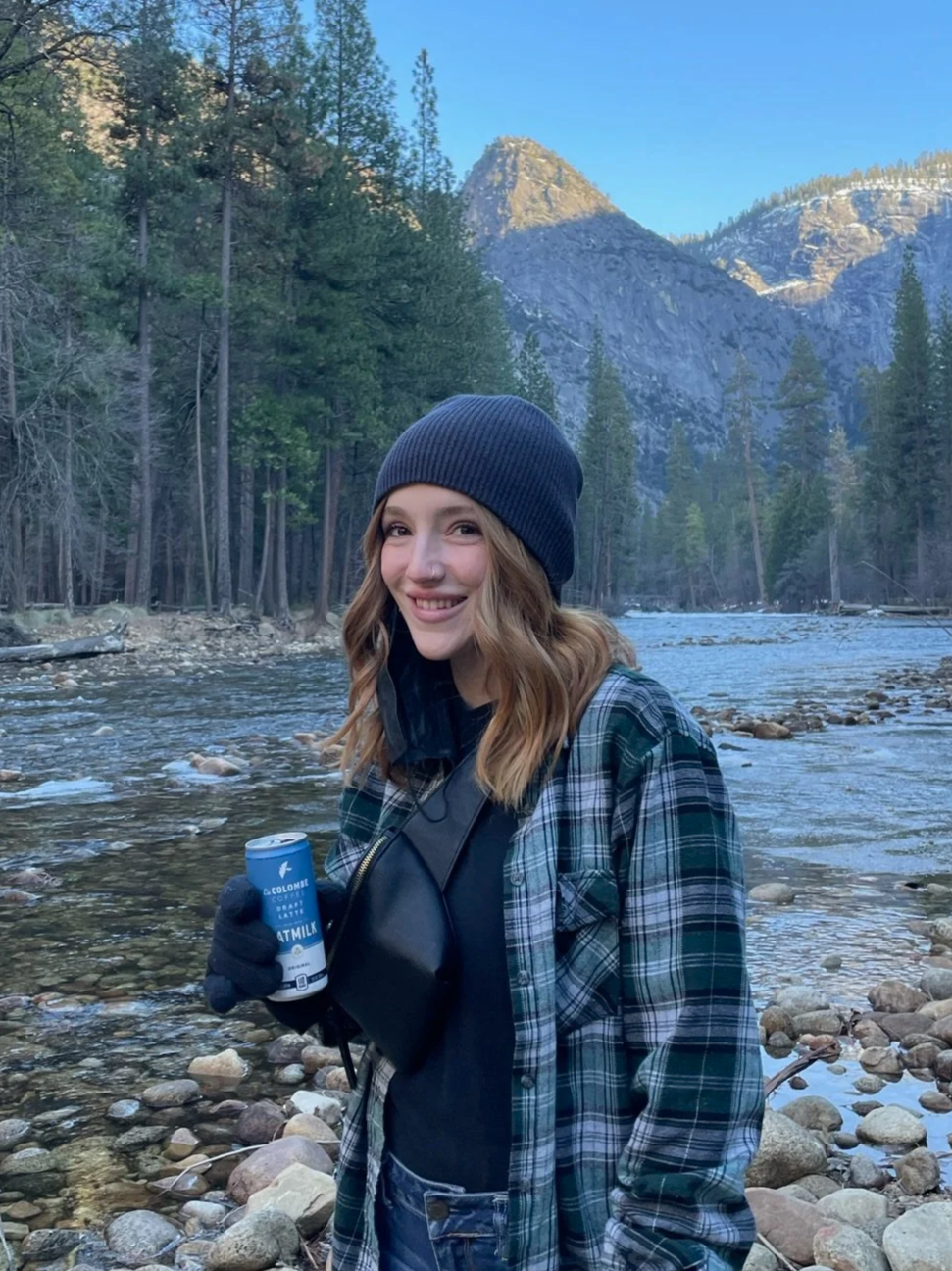A woman with wavy brown hair, wearing a dark beanie, black gloves, a plaid jacket, and a black shirt, standing by a rocky riverbank in a forested mountain landscape, holding a can of latte drink in her right hand.
