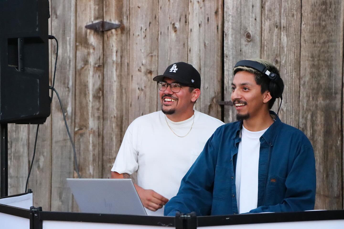 Two men, one with a baseball cap and glasses, and the other with headphones, stand in front of a rustic wooden wall, smiling and looking at a laptop.