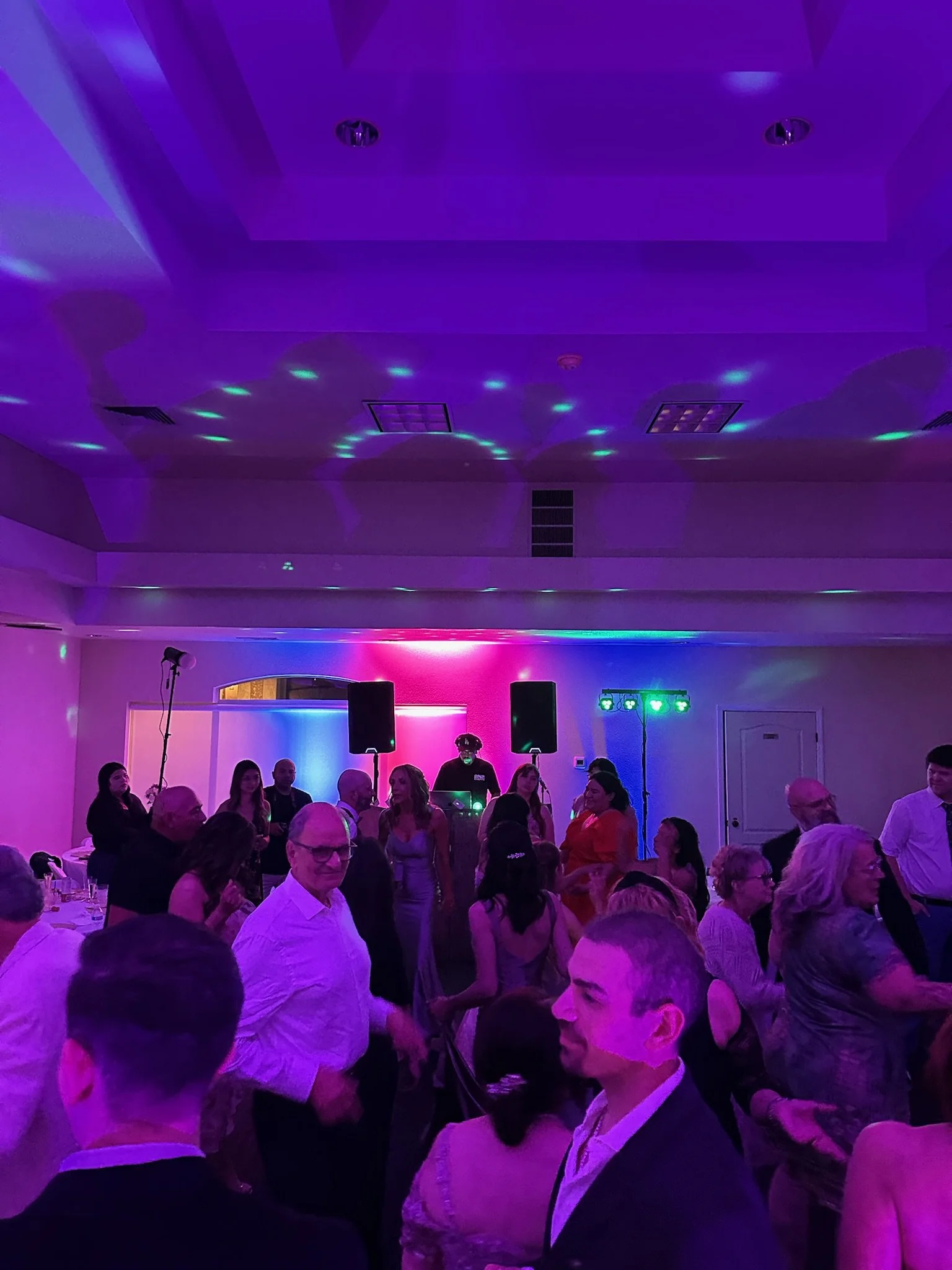 People dancing and socializing at a party or celebration in a dimly lit room with colorful purple and blue lighting.