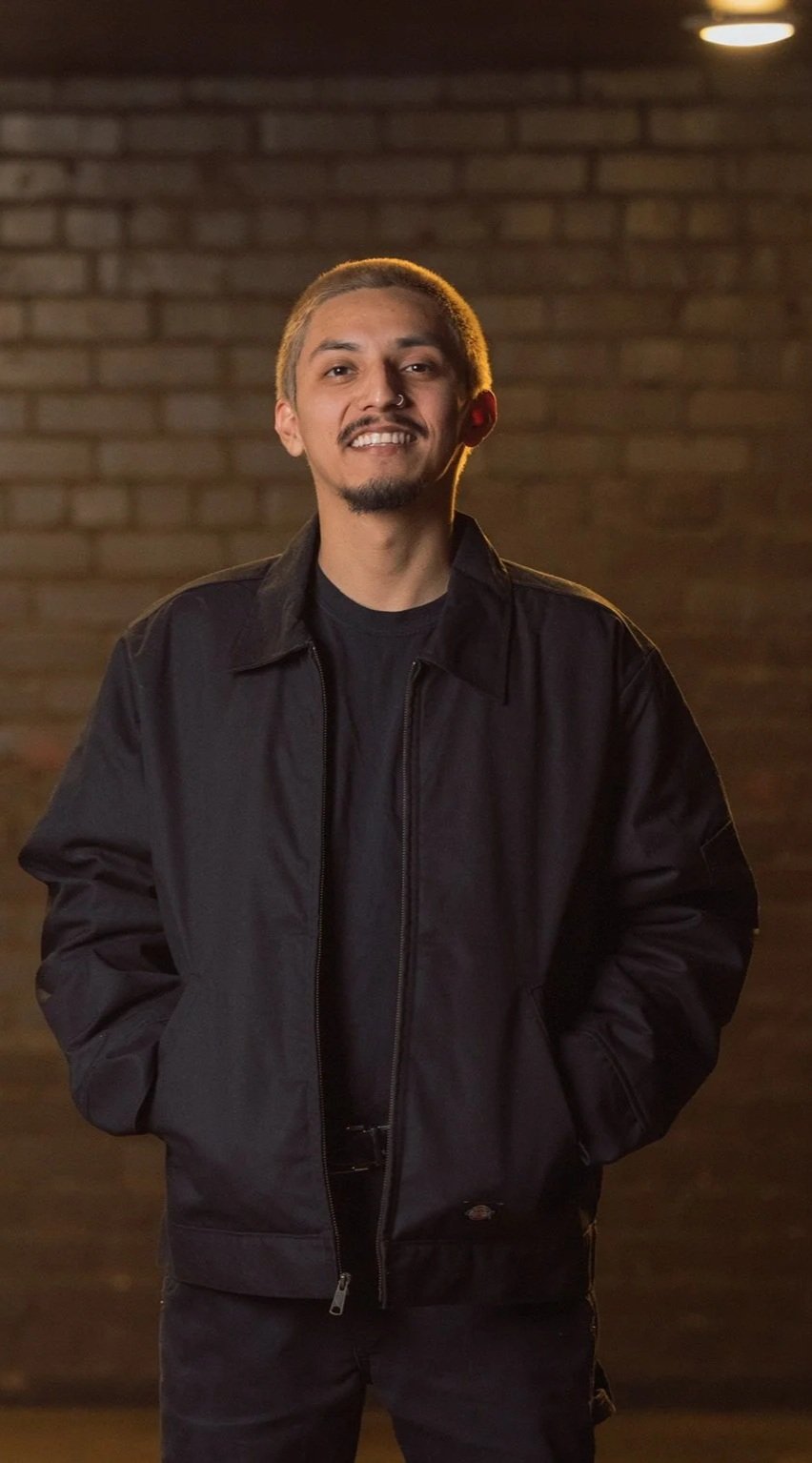 A smiling man with short hair and a mustache, wearing a black jacket and black t-shirt, standing in front of a brick wall at night.
