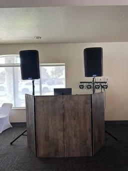 DJ equipment setup with two large speakers, a DJ controller, and a laptop on a wooden table in a room with large windows.