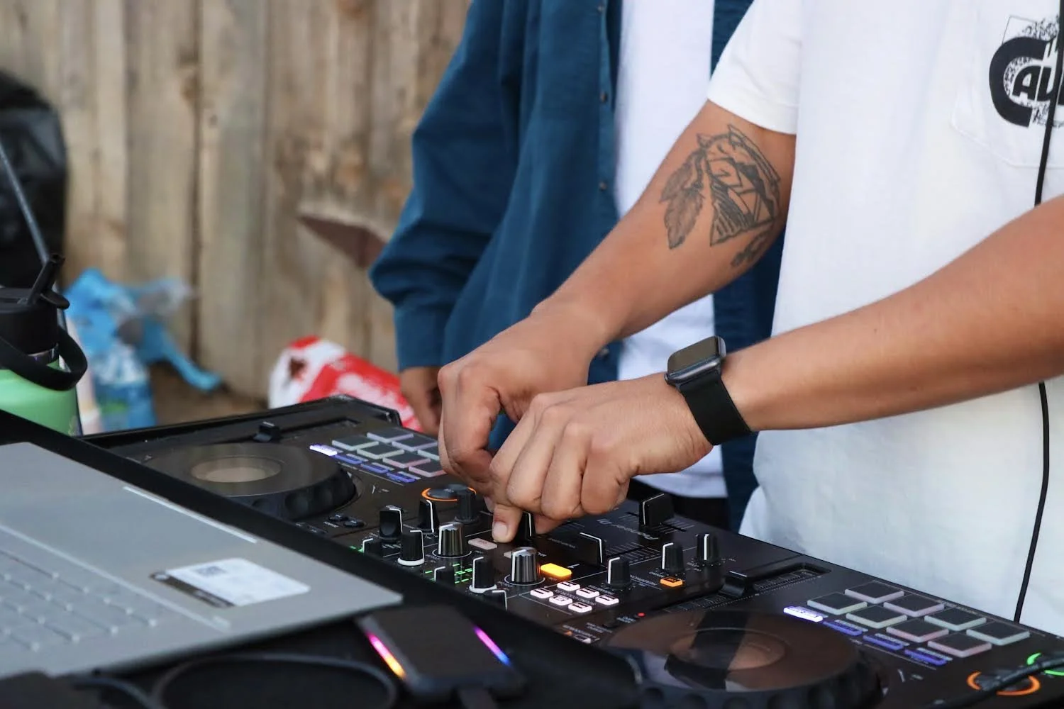 Close-up of a DJ's hands adjusting controls on a DJ mixer and a laptop, with another person partially visible in the background, all set against a rustic wooden backdrop.