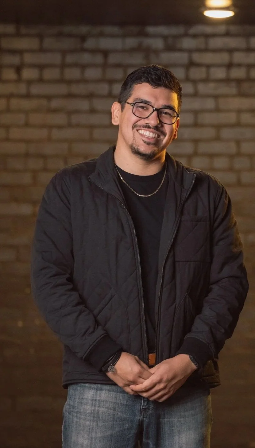 A smiling man with glasses, dark hair, and facial hair, wearing a black jacket, black shirt, and jeans, standing in front of a brick wall.