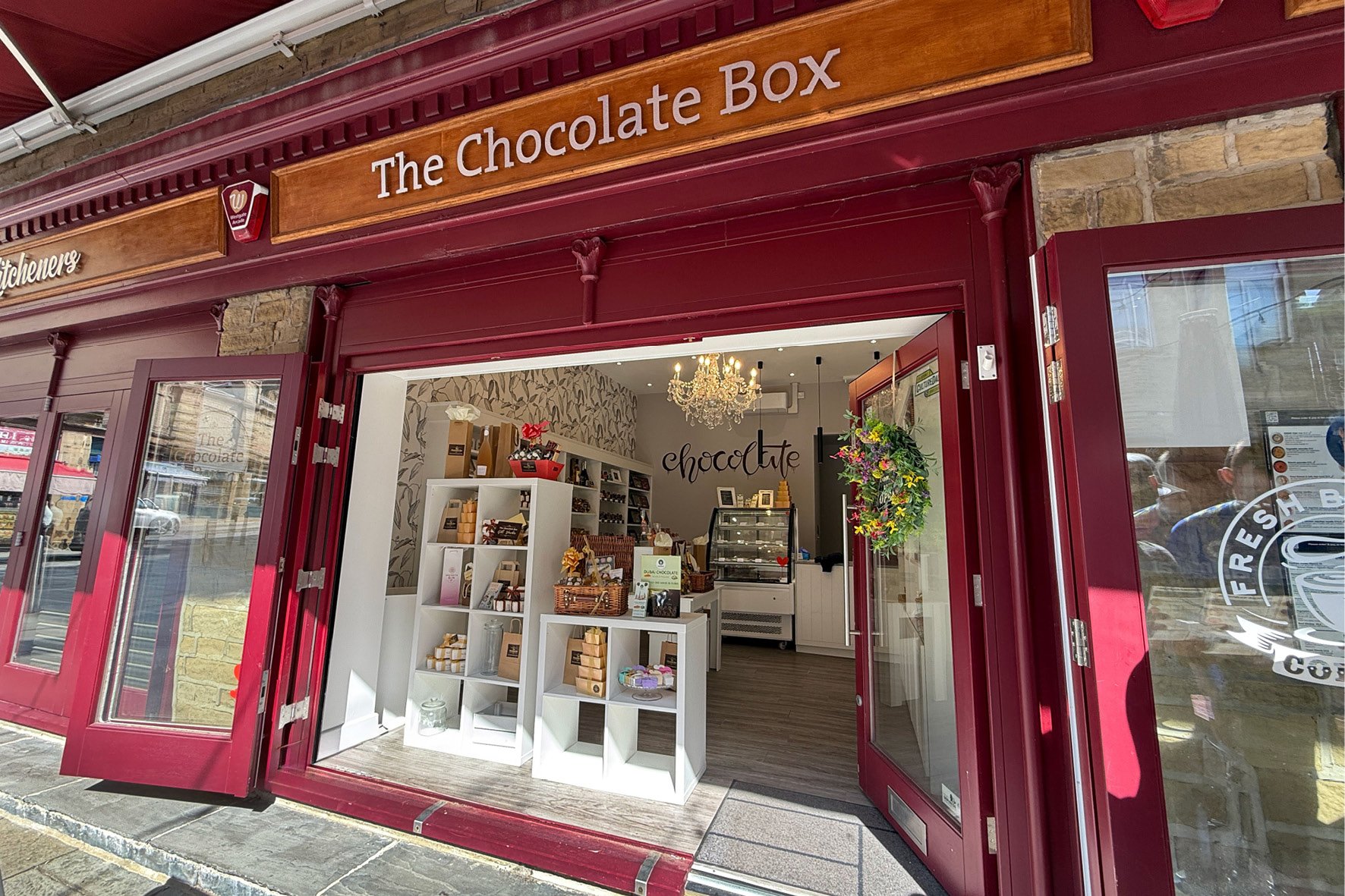 Exterior of a chocolate shop named 'The Chocolate Box' with open doors revealing a well-lit interior decorated with a chandelier, white shelves displaying chocolates and gifts, and a floral wreath hanging on the door.