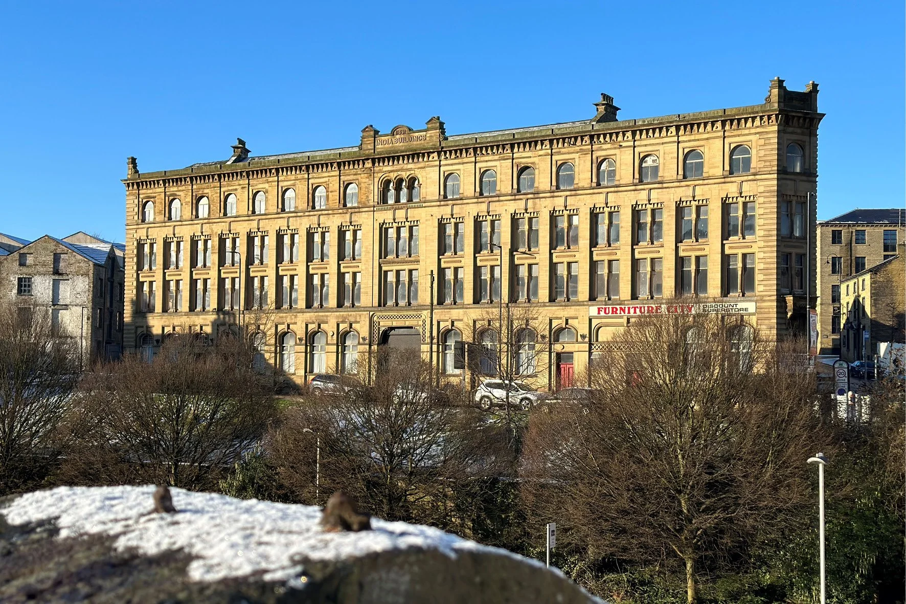Historic four-story yellow brick building with arched windows, a sign reading 'INDIA BUILDINGS' at the top, and a red and white sign for furniture store on the ground floor, viewed against a clear blue sky, with leafless trees and cars in the foreground.