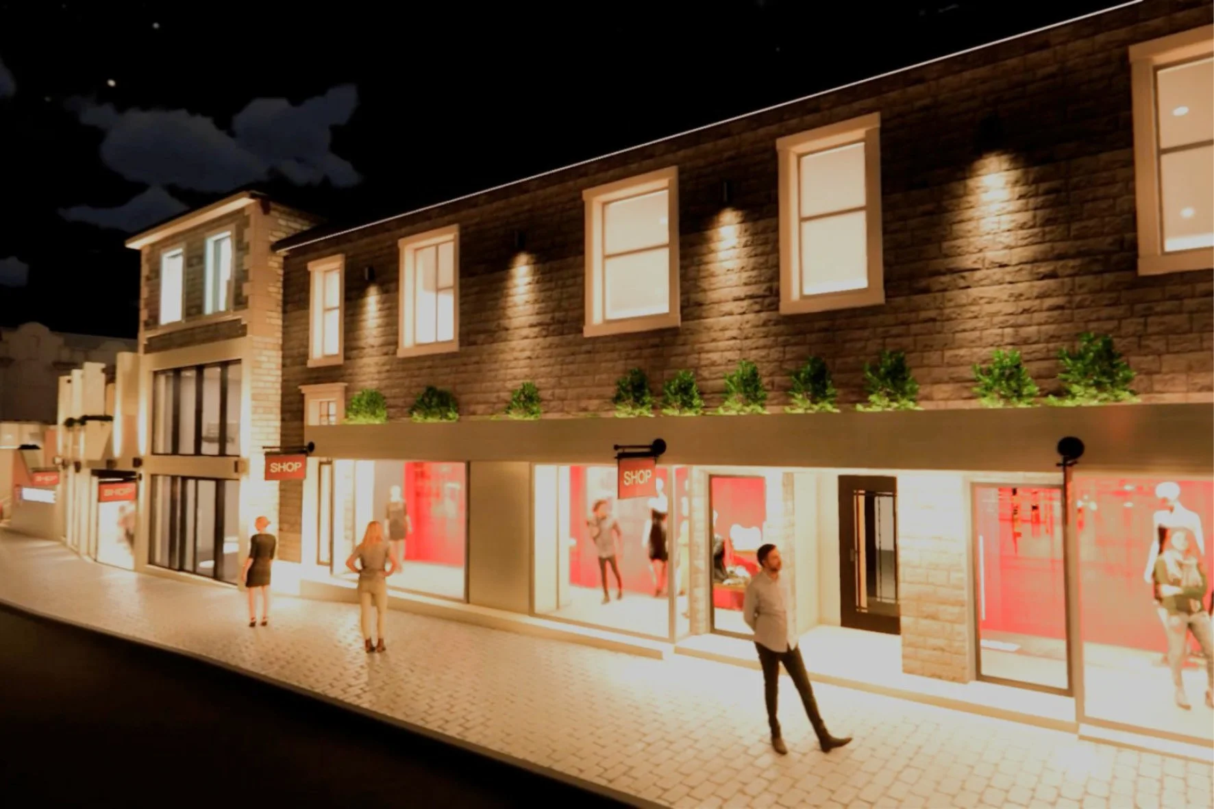 Night view of a modern shopping center with illuminated storefronts, people walking on the sidewalk, and an upper level with windows and green plants.