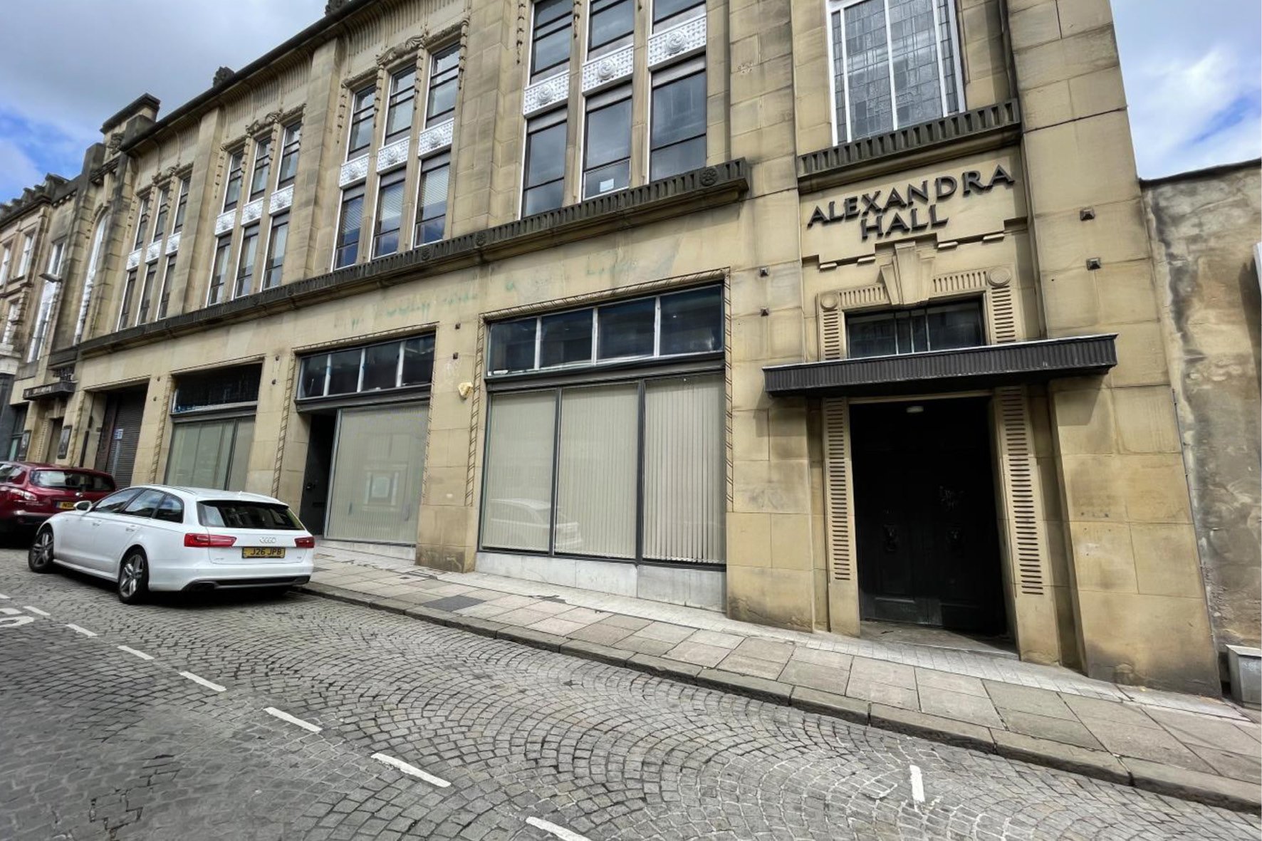 Facade of a historic building with an entrance labeled 'Alexandra Hall,' large windows, and stone exterior, situated on a cobblestone street with parked cars.