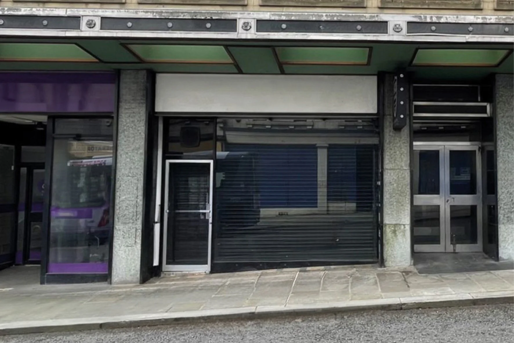 Empty storefront with closed black roller shutter, glass door, and surrounding windows, with sidewalk in front.