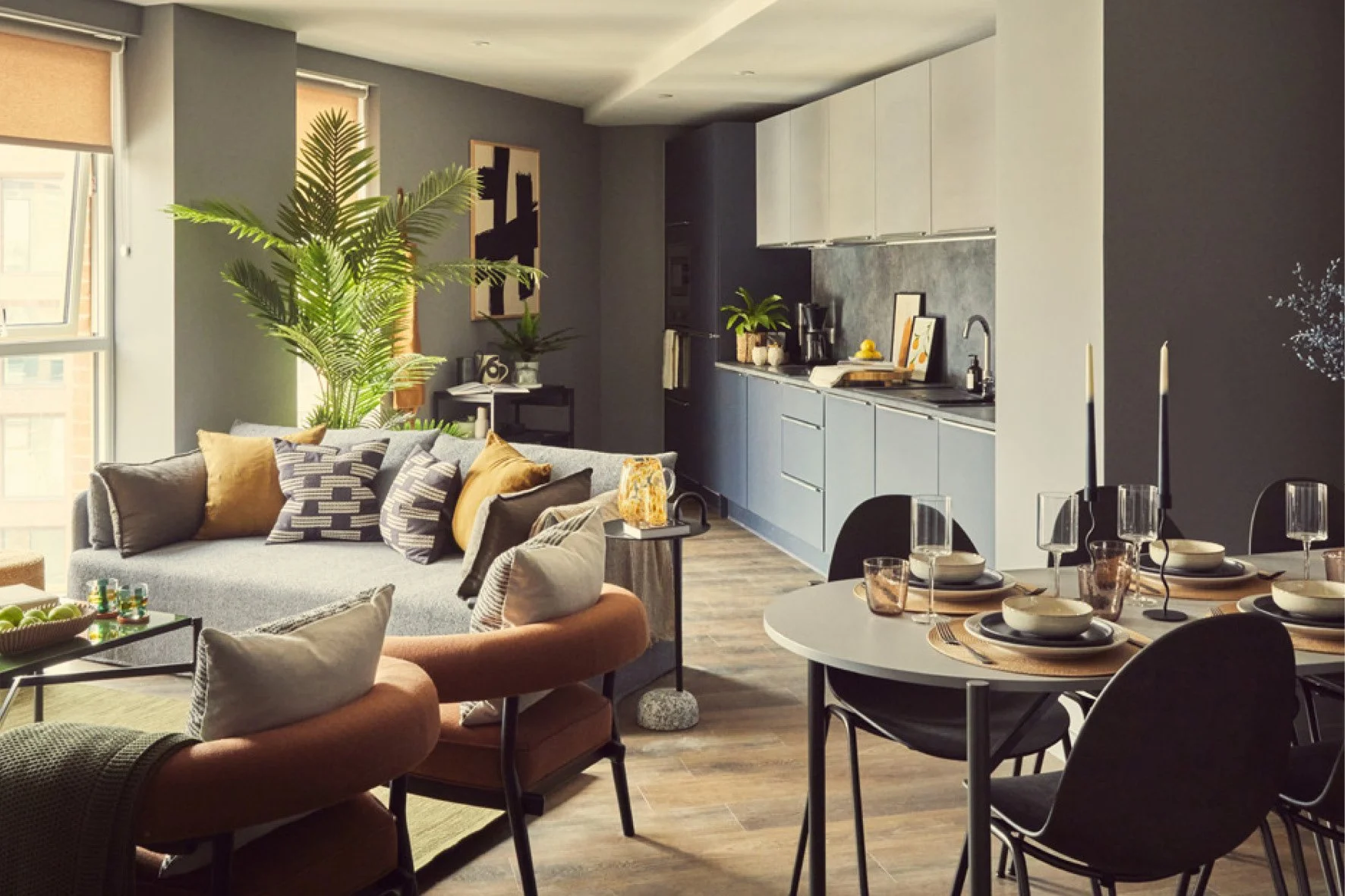 Modern apartment living room and dining area with gray and blue tones, large windows, a gray sofa with colorful cushions, a round dining table set for four, and a kitchen with light blue cabinets and black backsplash.
