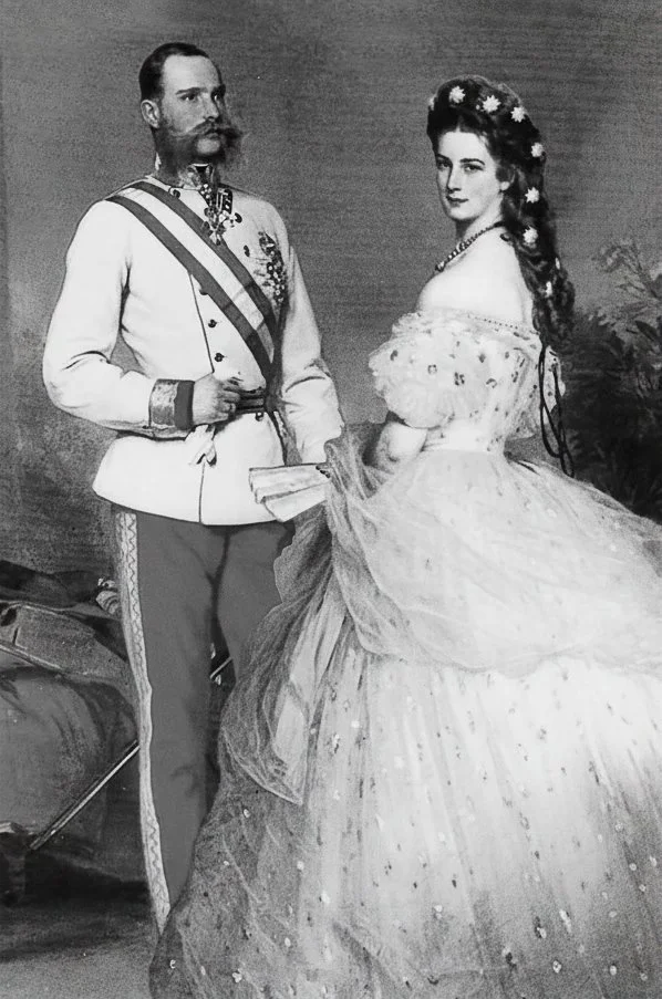 A black and white photograph of a man in a military uniform and a woman in a fancy dress, resembling royalty or aristocracy from a historical period, standing side by side indoors, with some plants and equipment in the background.