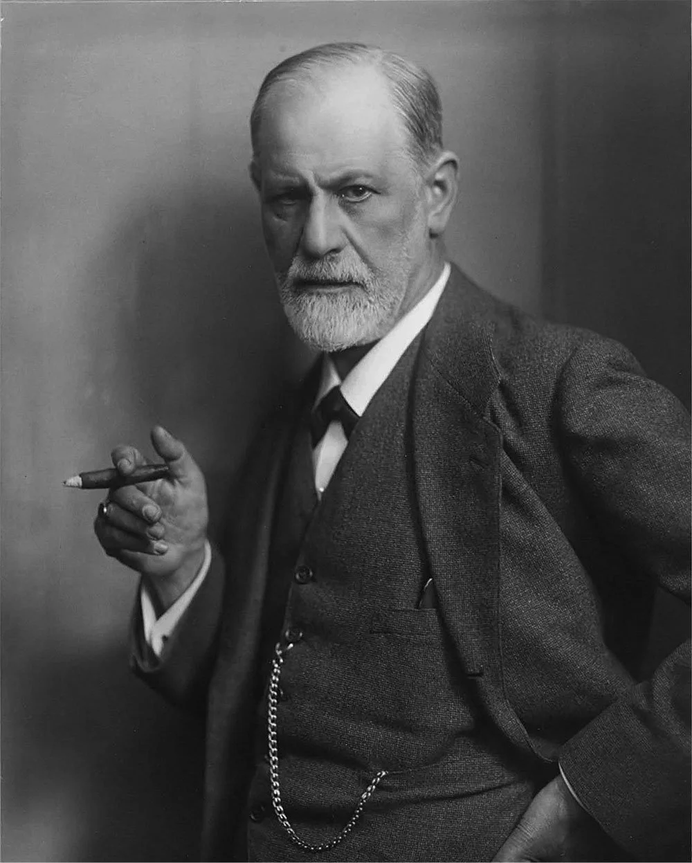 Black and white photograph of an older man with a beard and mustache, dressed in a suit, holding a cigarette in his right hand, and looking at the camera with a serious expression.