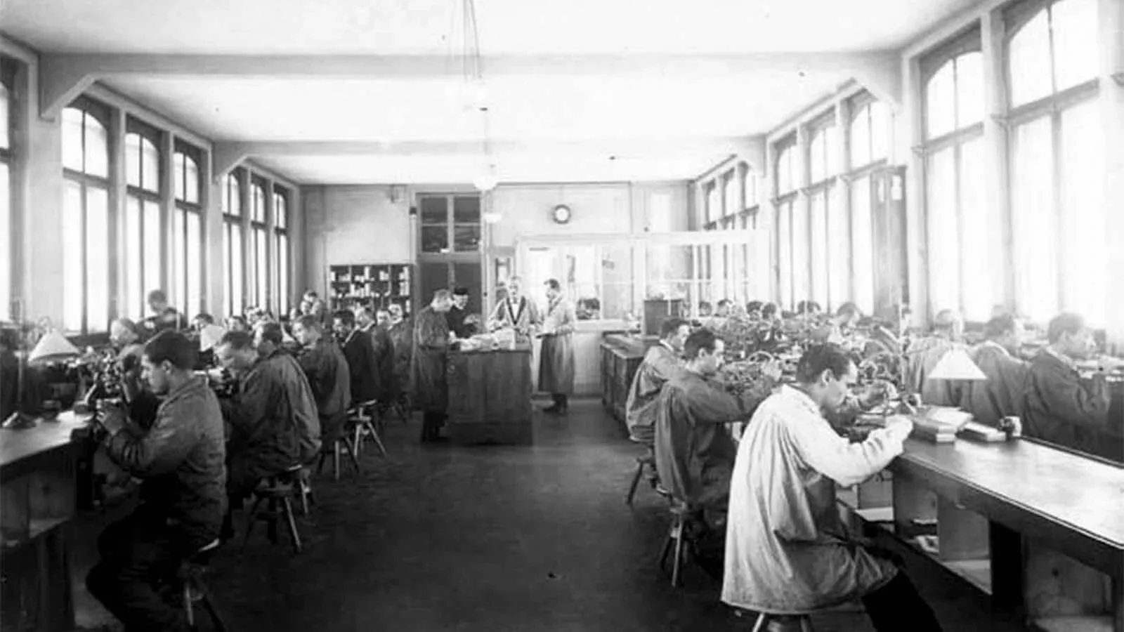 Black and white photo of students in a classroom with large windows, working at desks, some using microscopes and others reading or writing.
