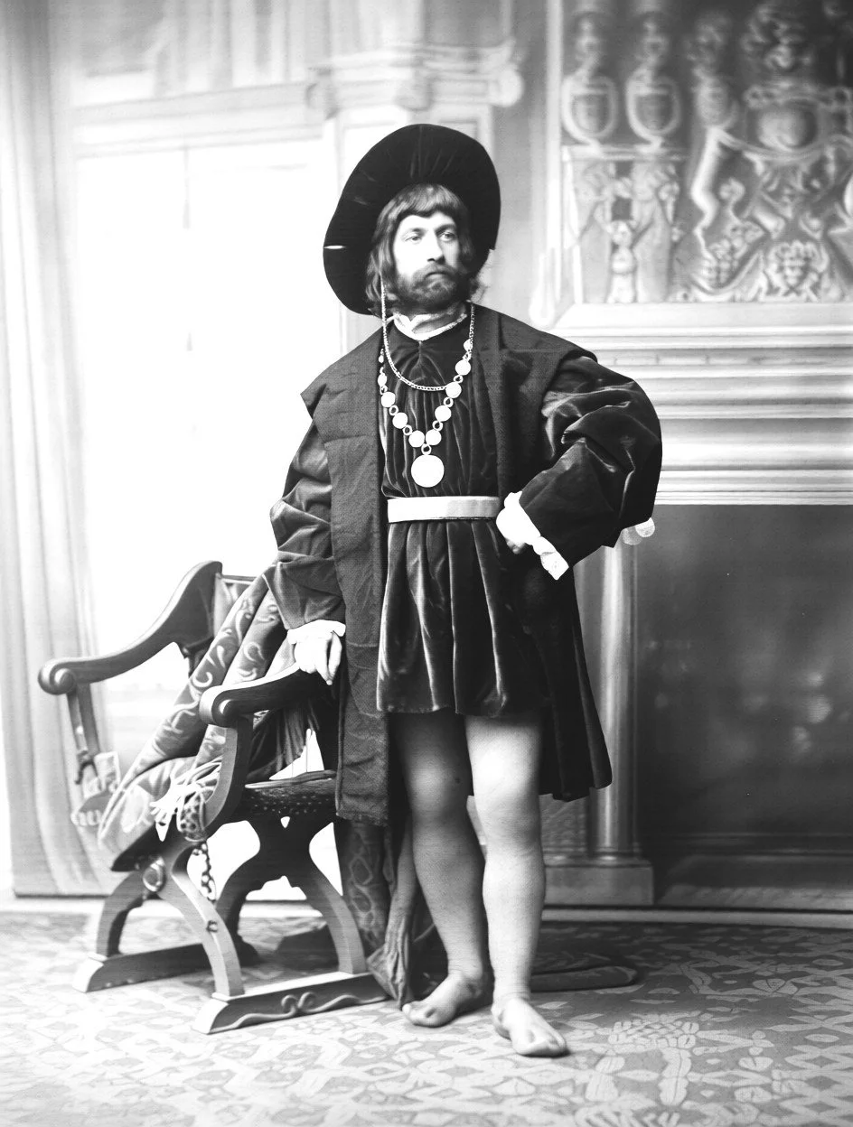 Black and white photo of a man in historical costume with jewelry, a wide-brimmed hat, and stockings, standing indoors with an ornate wooden chair and detailed wall decor in the background.
