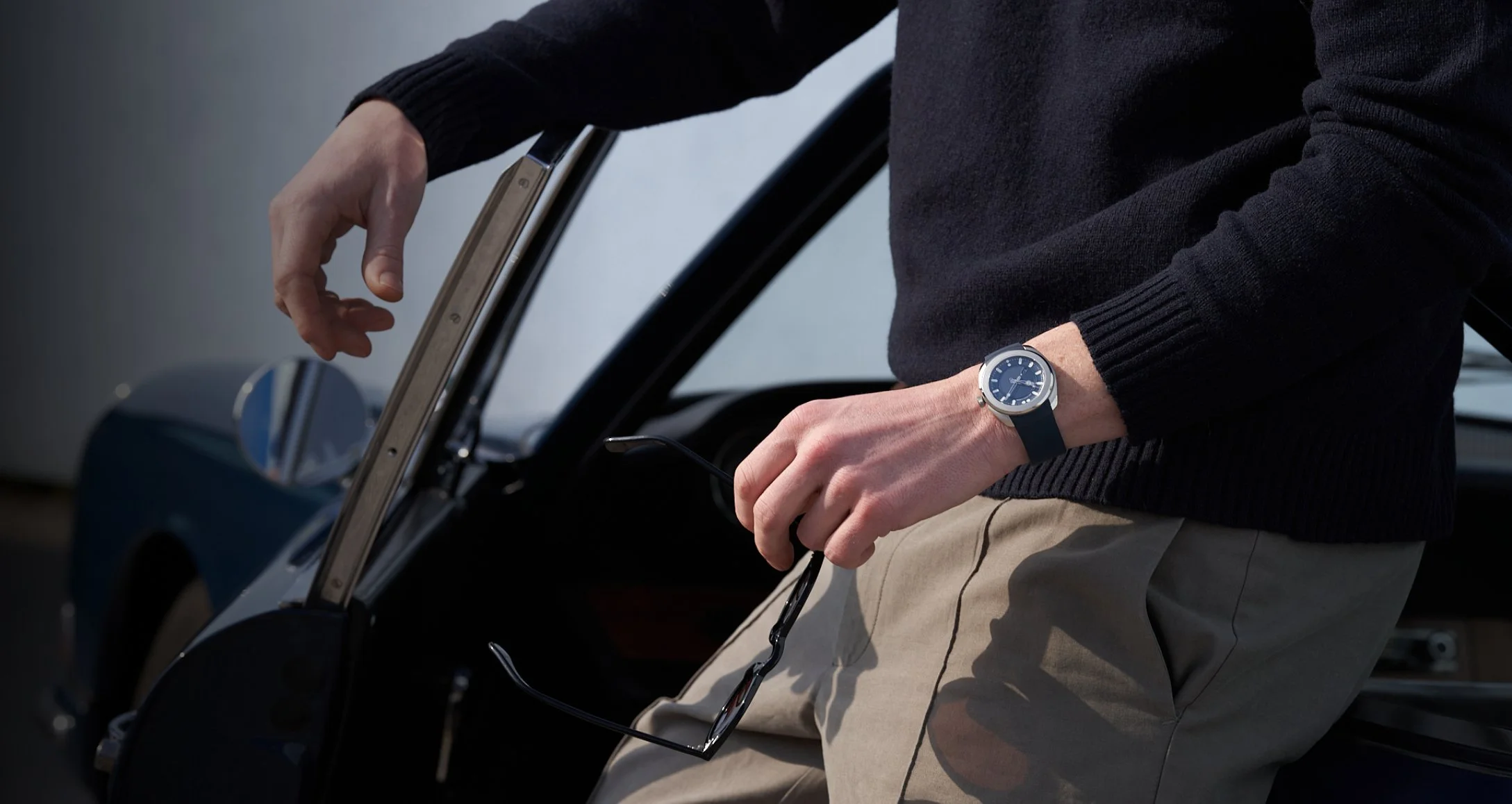 Close-up of a man wearing a black sweater and beige pants, holding eyeglasses in one hand and a smartphone in the other, standing near a black car. The man is wearing a silver wristwatch with a blue face and black strap.