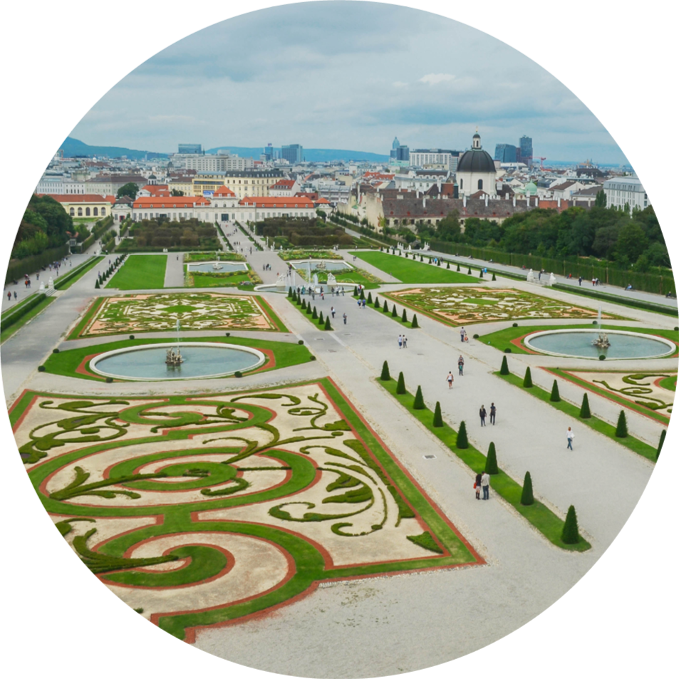 A large, formal garden with intricate flower beds, fountains, and walks, set against a cityscape with historic and modern buildings under a cloudy sky.