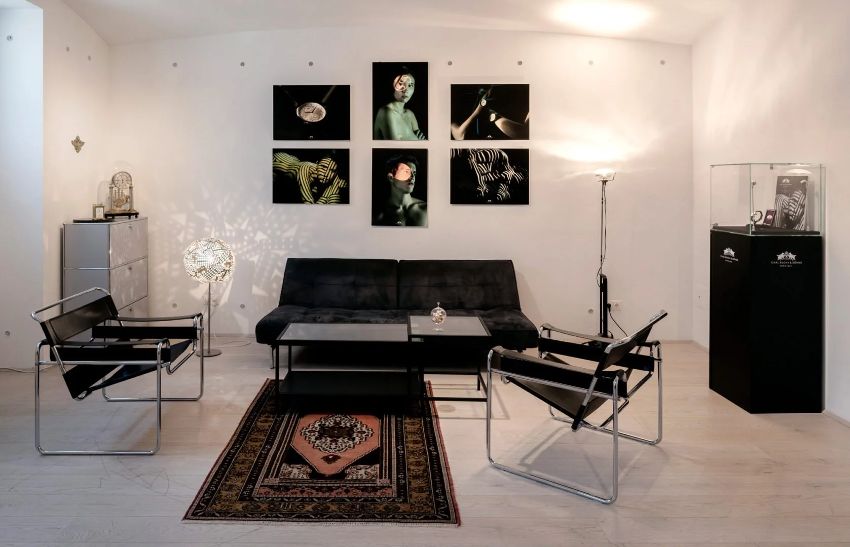 Modern living room with black sofa, two black armchairs, glass-topped coffee table, decorative lamp, artwork on the wall, and a display case with collectibles.