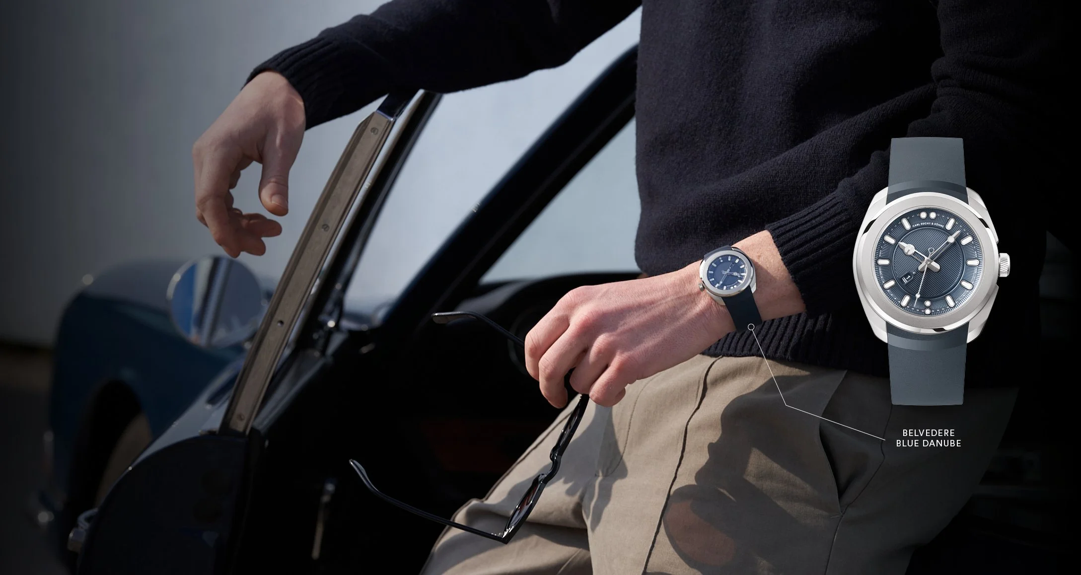 Person standing next to a car, wearing a black sweater, beige pants, and a Belvedere Blue Danube watch on their wrist, holding a pair of sunglasses and a smartphone.