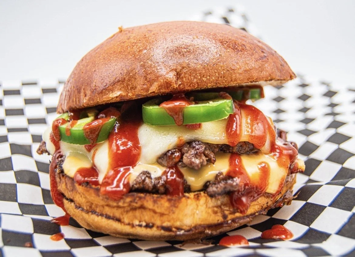 Close-up of a burger with cheddar cheese, beef patty, jalapenos, ketchup, and a toasted bun on checkered paper.