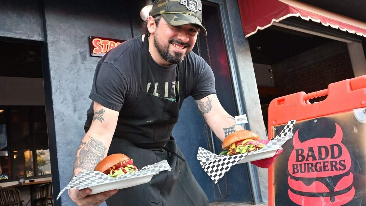 Man smiling and presenting two burgers on plates outside a restaurant called BADD BURGERS