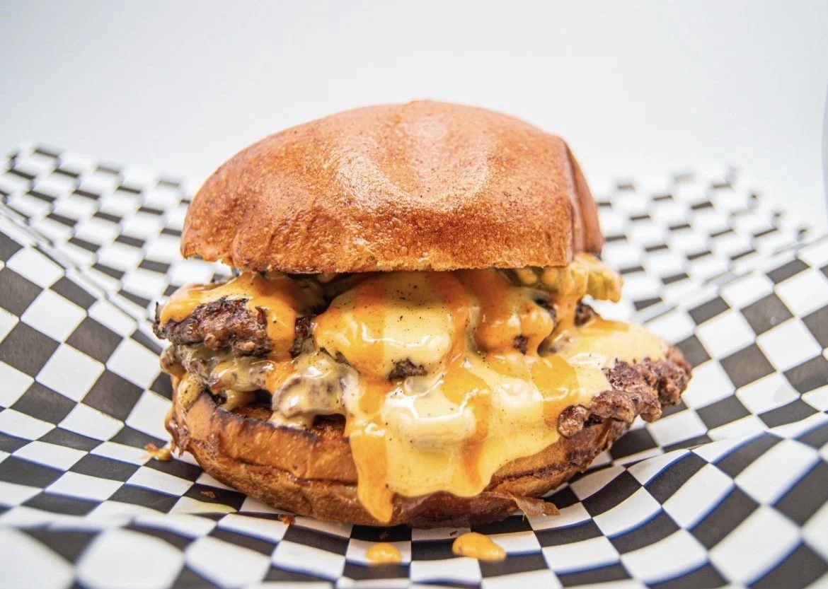 Close-up of the namesake The Badd Burger with melted American cheese, beef patties, pickles, Signature Badd Sauce between two brioche buns on checkered paper
