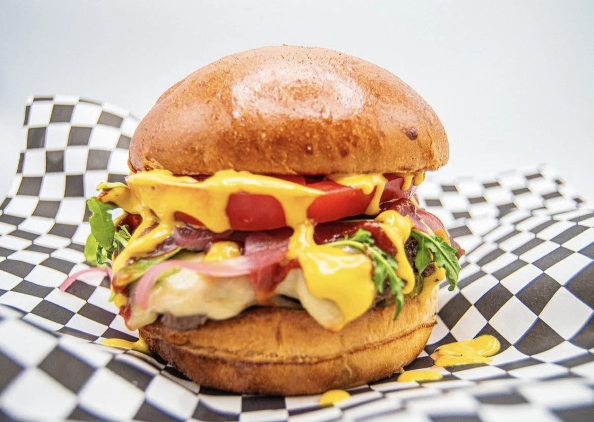 Close-up of a cheeseburger with lettuce, tomato, pickles, onions, cheese, and bacon in a bun, on checkered paper