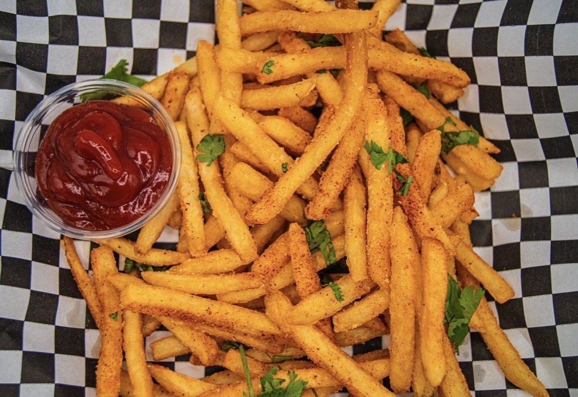 A basket of seasoned French fries garnished with chopped parsley and a small cup of ketchup on black and white checkered paper