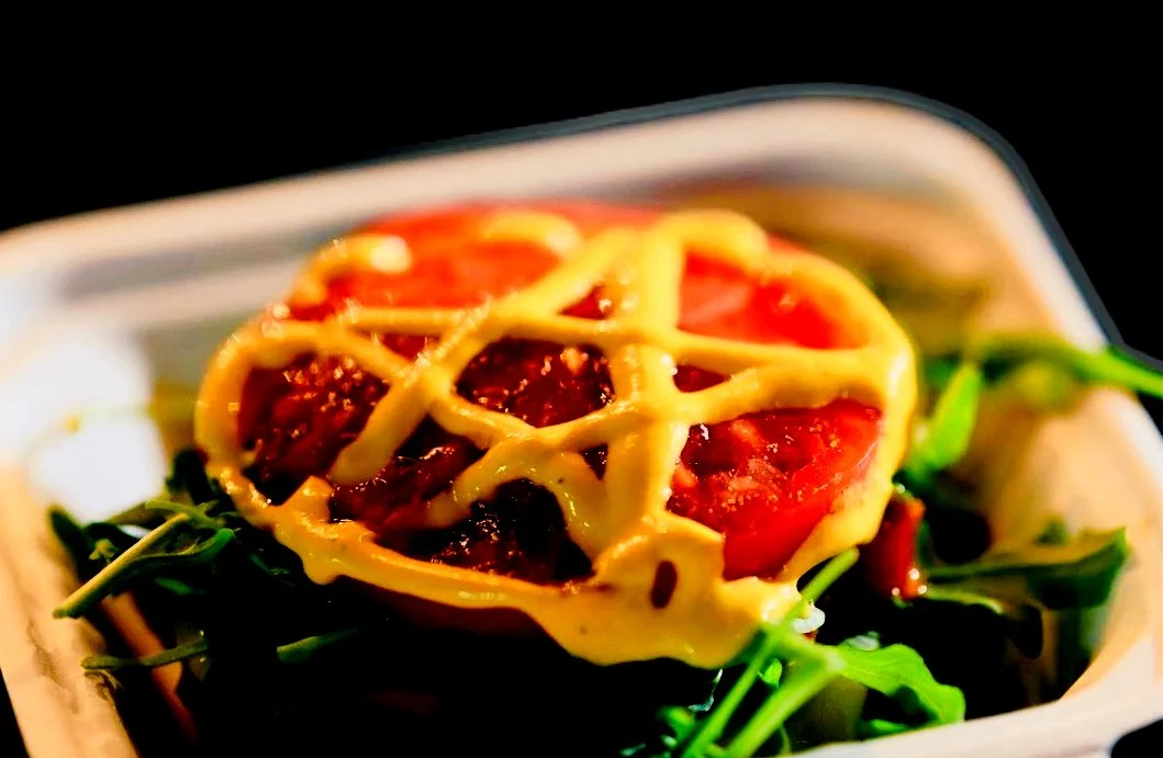Close-up of a small dish with a piece of baked pasta topped with ketchup and a net of melted cheese, served over fresh greens.