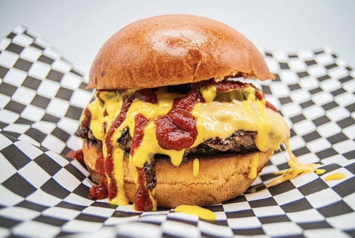 Close-up of a The McBaddy with melted American cheese, Badd Blood spicy ketchup, The Gold pickled mustard, pickles, and a toasted brioche buns on checkered paper