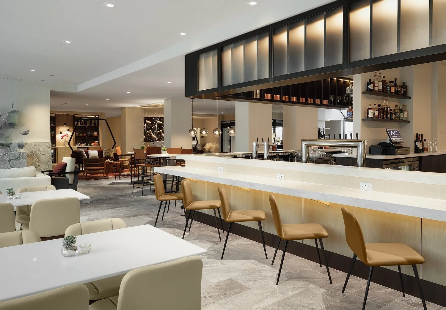 Modern hotel bar with tan chairs and white countertops, shelves with liquor bottles, and seating area in the background.
