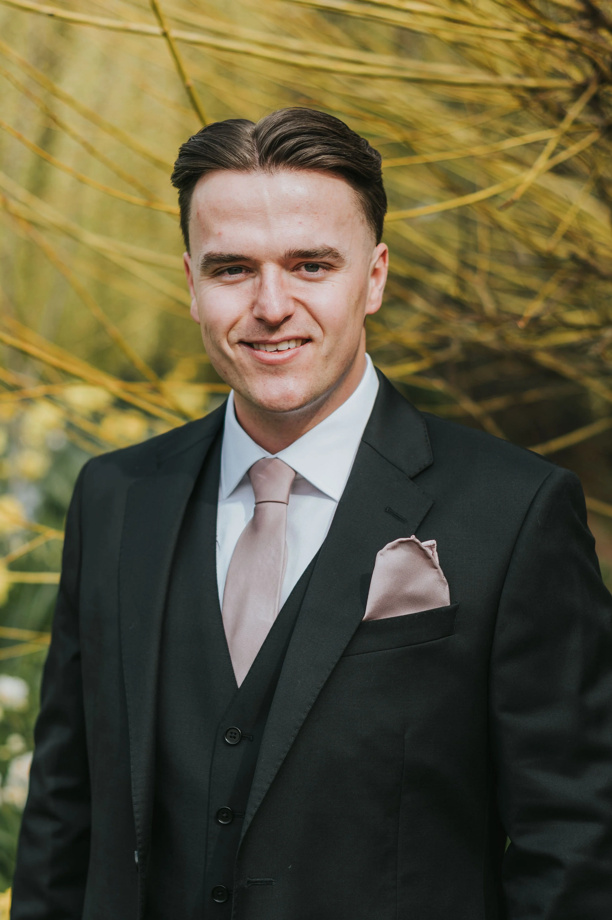 A young man in a black suit with a light pink tie and pocket square, smiling outdoors with yellowish-green foliage in the background.