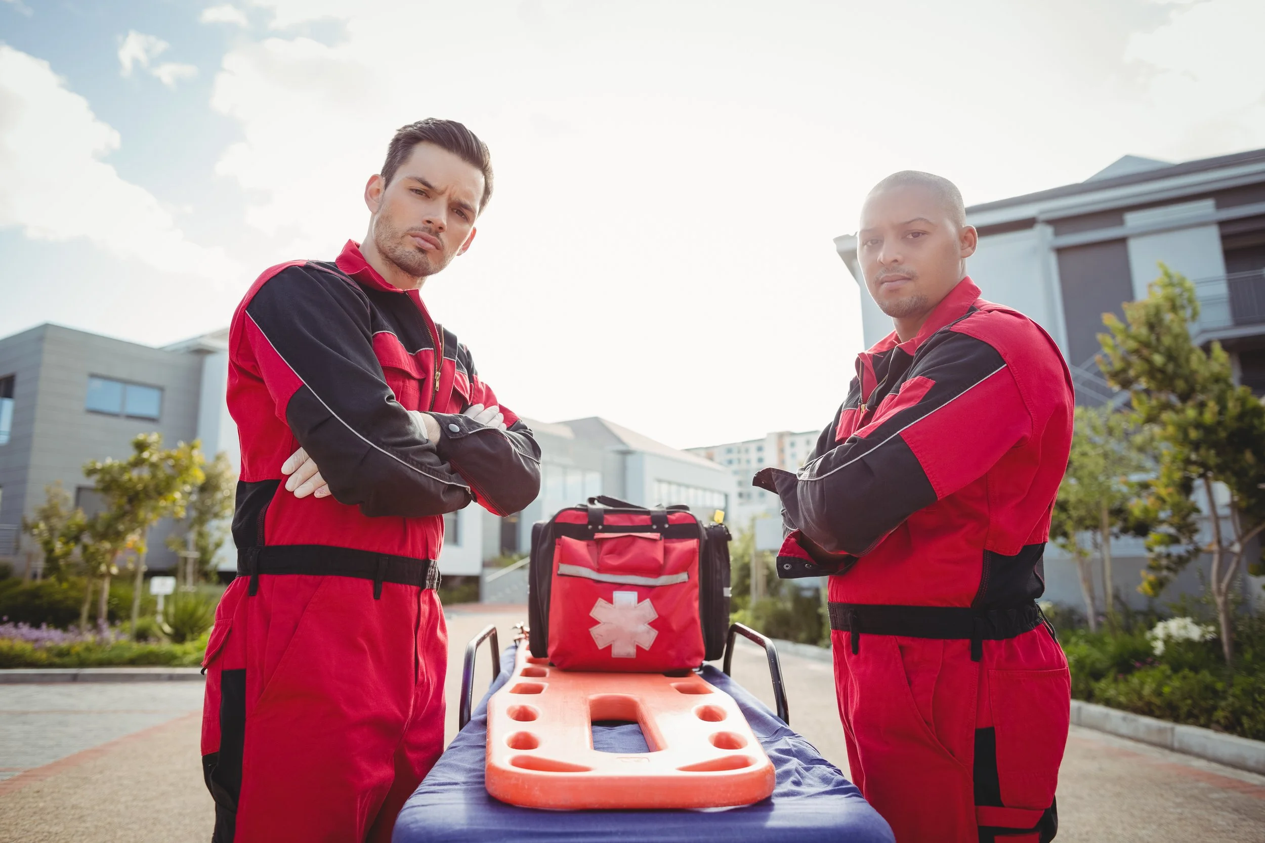 Two emergency medical responders wearing red and black uniforms standing outdoors with a stretcher and a red medical bag with a white cross. They look serious and alert.