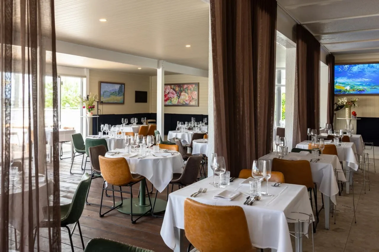 Elegant restaurant dining area with white tablecloths, set with wine glasses, water glasses, silverware, and napkins. Multiple colorful chairs surround the tables, with artwork on the walls and curtains dividing parts of the space.