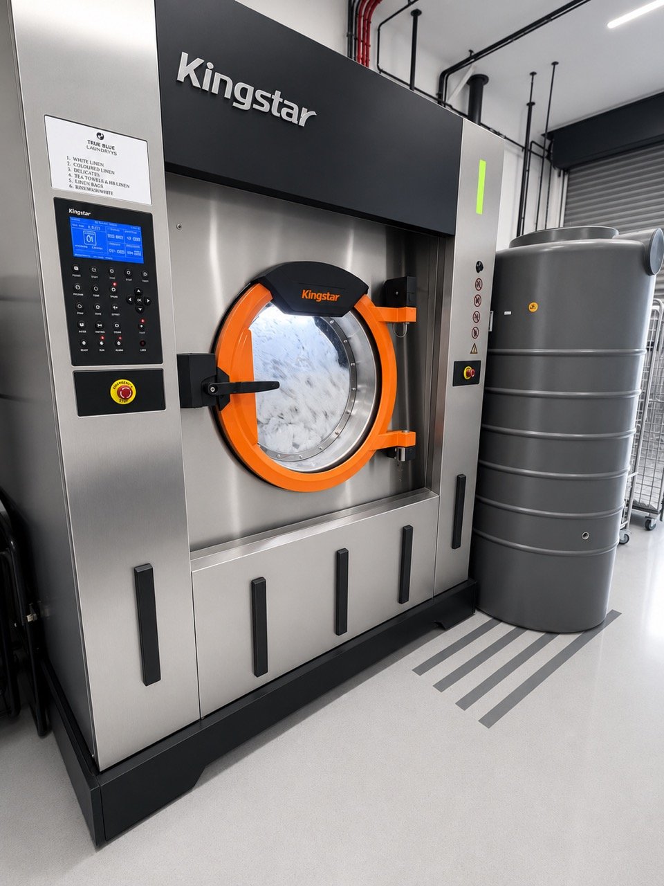 Commercial industrial washing machine by Kingstar with digital control panel and bright orange door handle, situated in a laundry or industrial facility with a gray water tank nearby.