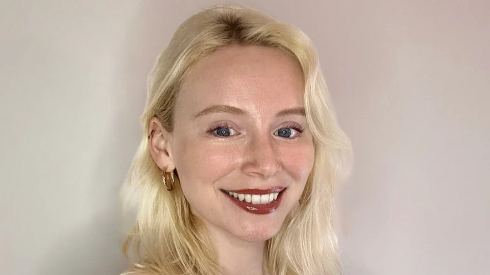 A woman with blonde wavy hair, fair skin, blue eyes, wearing gold hoop earrings, smiling against a plain light background.