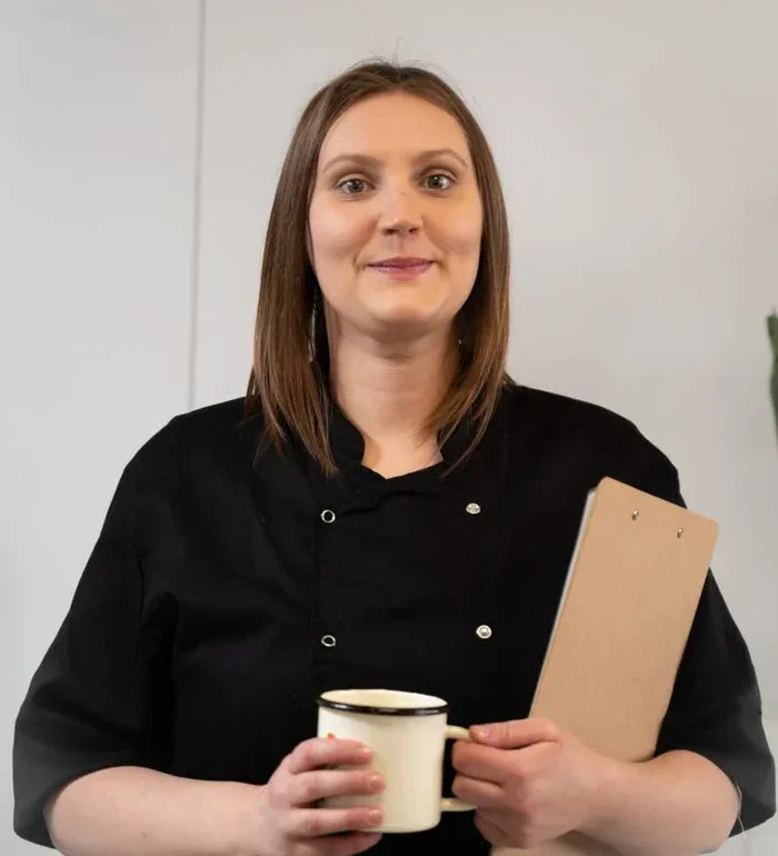 A woman with shoulder-length brown hair wearing a black chef's coat holding a mug and a clipboard, standing indoors.