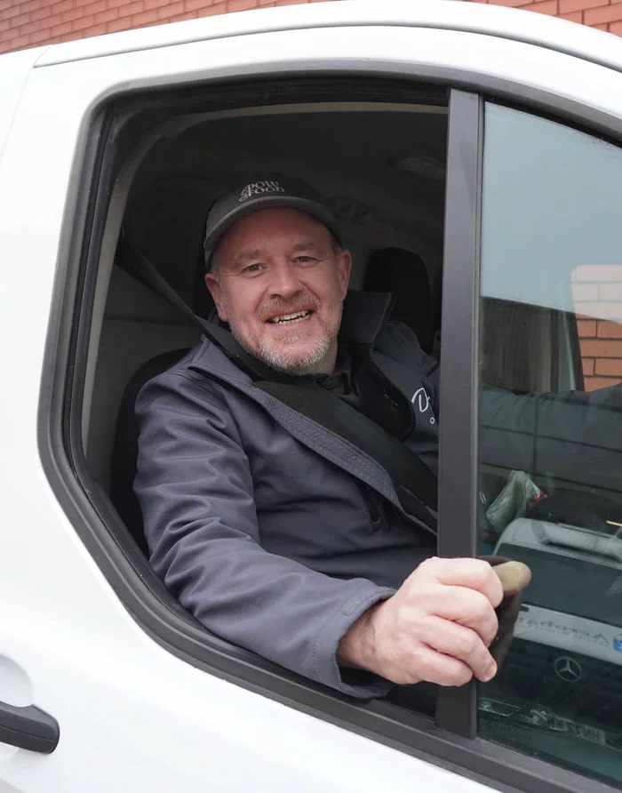 A smiling man with a gray beard and mustache inside a white vehicle, wearing a dark jacket and a dark baseball cap, looking out the driver's window.