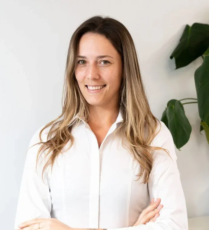 Smiling woman with long, wavy brown hair wearing a white button-up shirt, standing in front of a white wall with green plant leaves on her right.