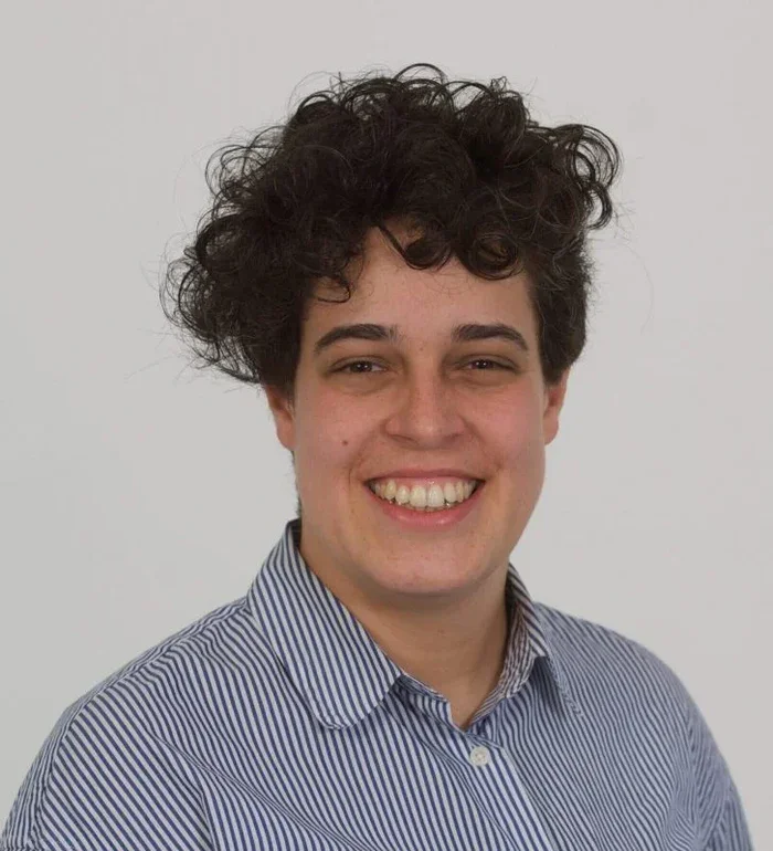 A young person with dark, curly hair and fair skin, smiling, wearing a blue and white striped collared shirt, against a plain light gray background.