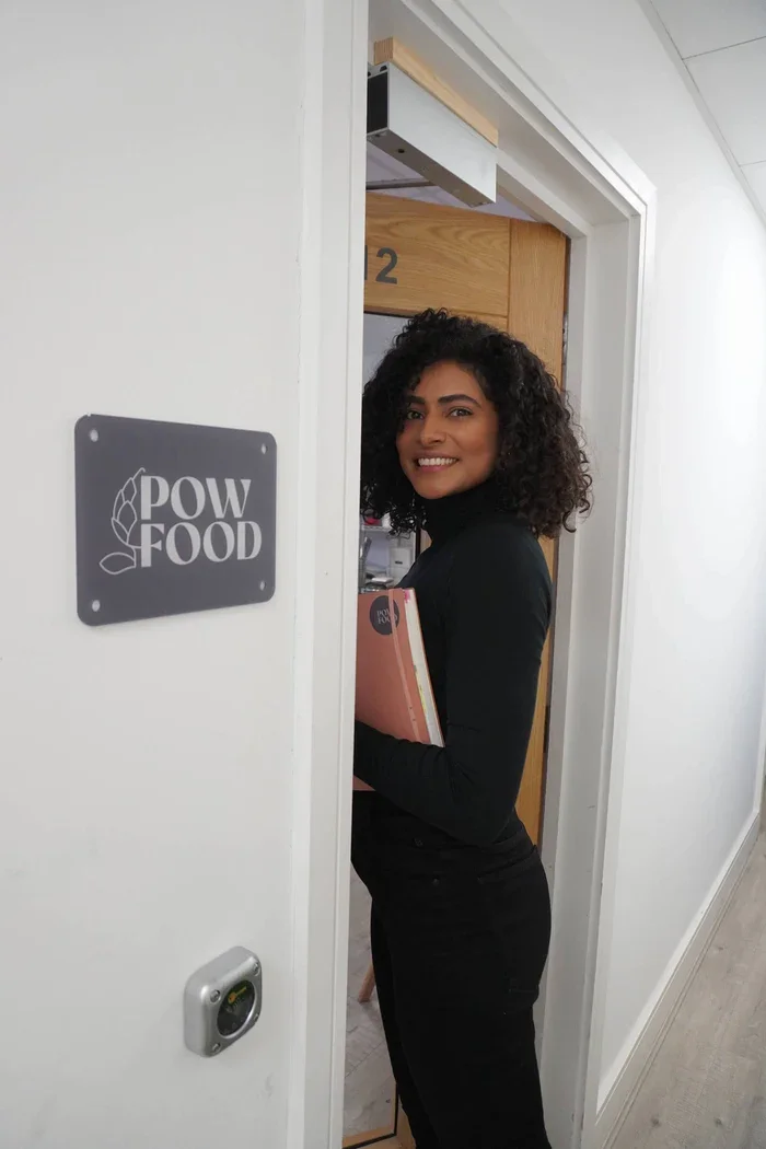 Woman with curly hair smiling and holding a pink notebook, standing in a doorway labeled '2' outside a room, next to a sign that says 'POW FOOD'.