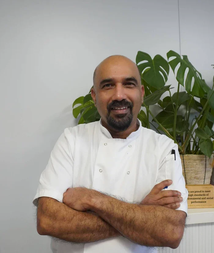 A man in a white chef's uniform standing with arms crossed in front of a large potted plant indoors.
