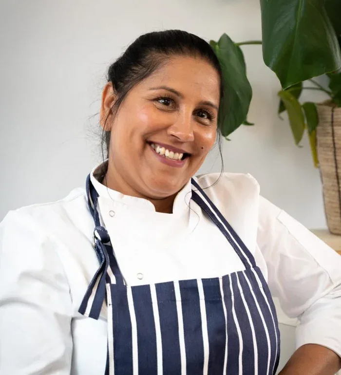 A smiling female chef with dark hair tied back, wearing a white chef's coat and a navy blue and white striped apron, sitting in a kitchen with a large potted plant in the background.