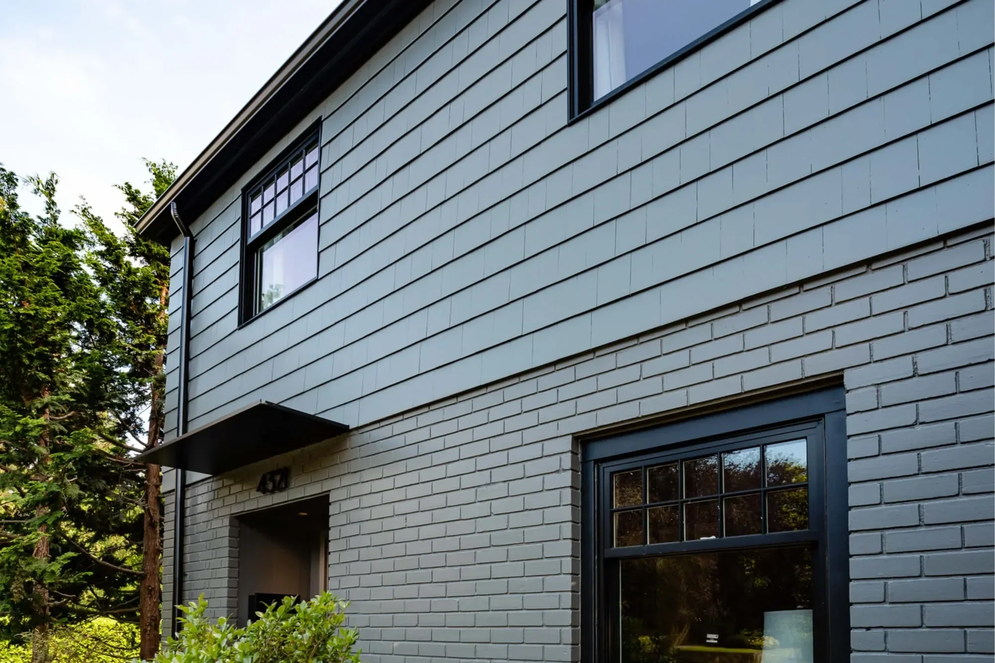 CBG CONSTRUCTION SOLUTIONS PTY LTD BEST COMPANY IN MELBOURNE Close-up view of a modern, two-story house with gray brick and horizontal siding exterior, black framed windows, and greenery in front.