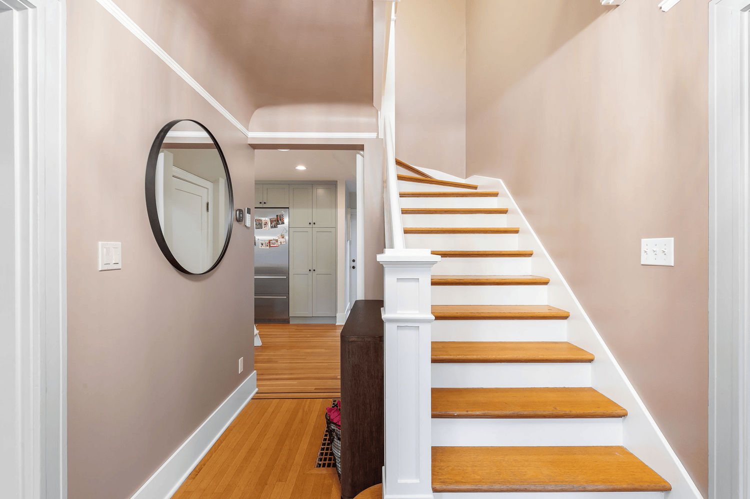 CBG CONSTRUCTION SOLUTIONS PTY LTD BEST COMPANY IN MELBOURNE Interior view of a house entrance with a staircase on the right, a mirror and light switch on the beige wall to the left a kitchen with closed cabinets and a refrigerator in the background.